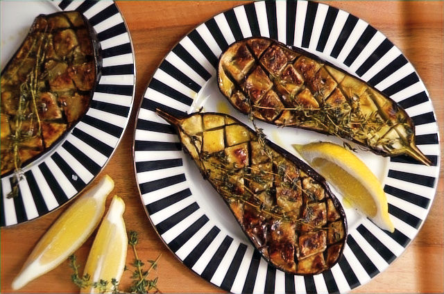 Grilled eggplant halves with thyme sprigs on a black and white striped plate; lemon wedges on the side. Rustic wood background, vibrant and appetising presentation.