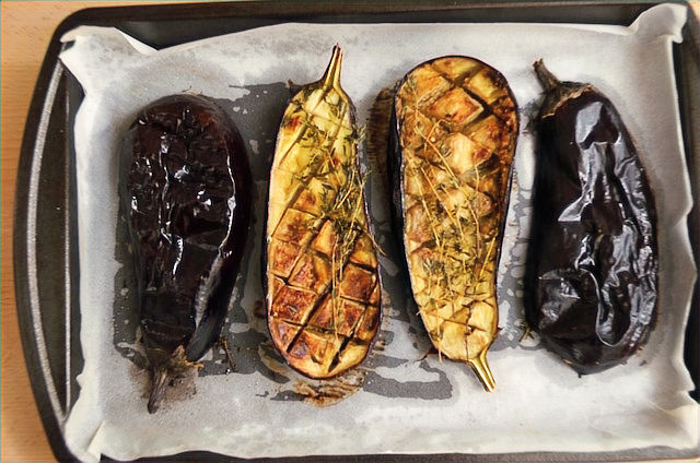 Four roasted eggplant halves on a baking sheet lined with parchment paper; two are halved with crisscross cuts. Rustic kitchen vibe.