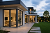 Casa contemporanea con grandi vetrate dai profili scuri, terrazza e giardino illuminato al crepuscolo.