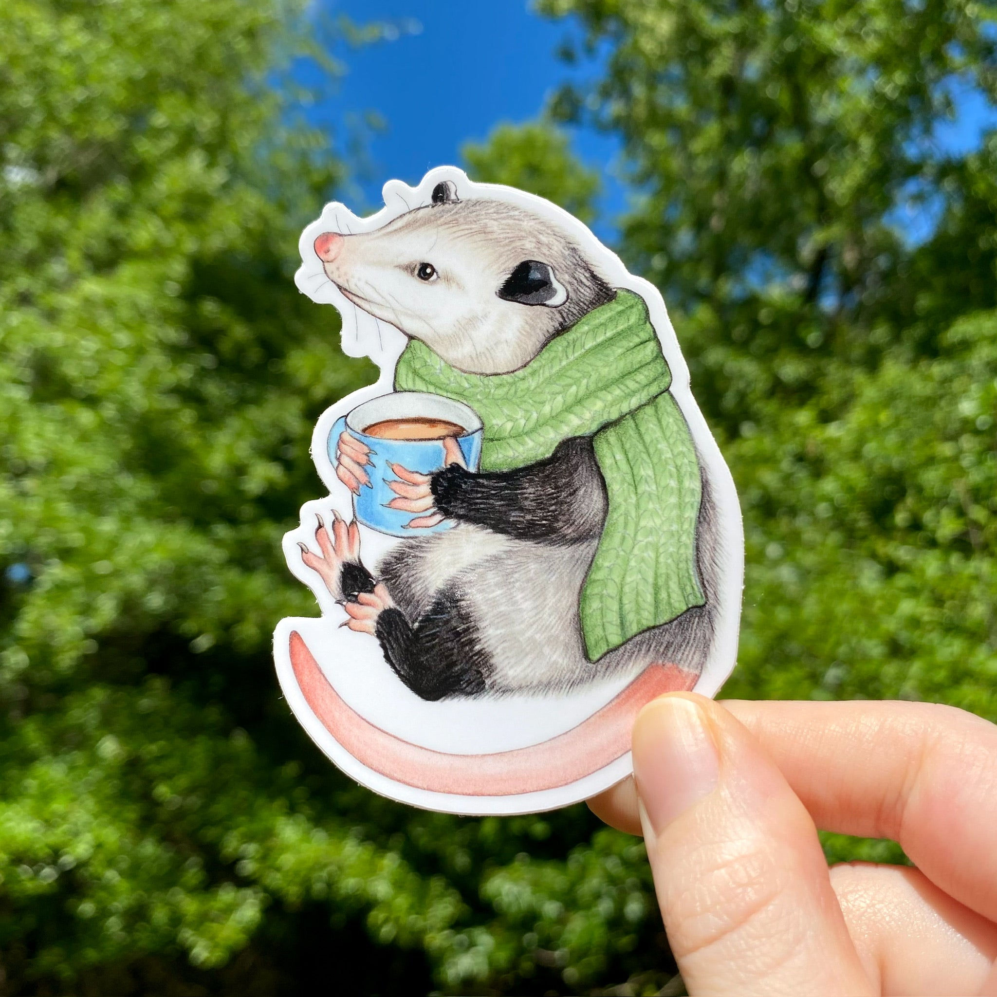 A hand holding a sticker of an opossum and a scarf holding a mug of hot chocolate
