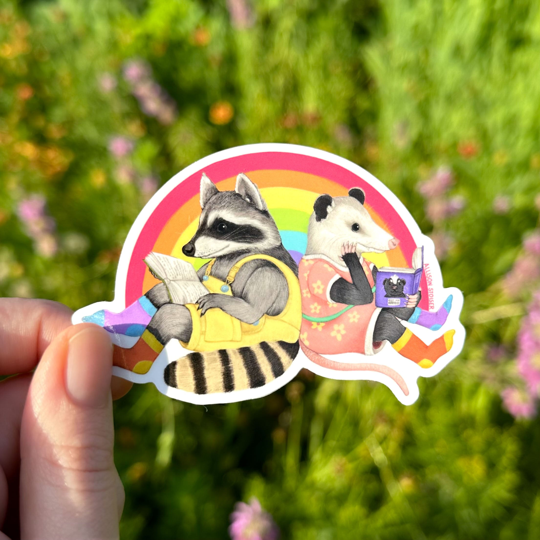 a hand holding a sticker of a raccoon and opossum sitting back to back and reading in front of a rai