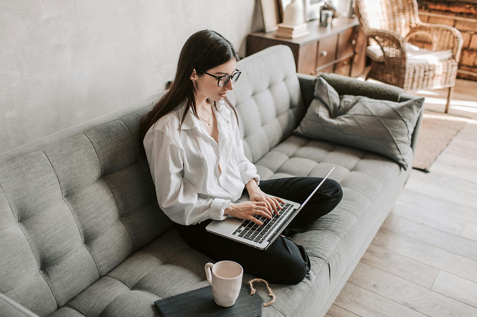 Seorang perempuan sedang duduk bersila di atas sofa, sebuah laptop terbuka di pangkuannya