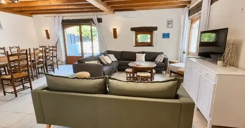 Intérieur spacieux du gîte Les Tournesols montrant un grand canapé d'angle bleu, une télévision, une table en bois et des éléments en pierre traditionnelle.
