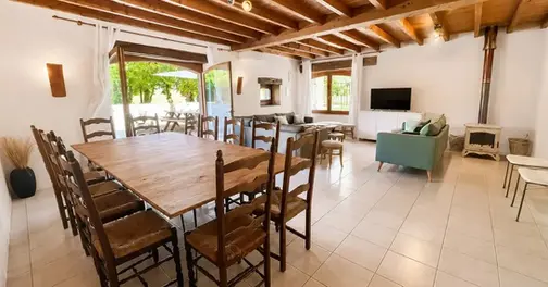 Pièce de vie lumineuse du gîte Les Tournesols avec grande table en bois pour 12 personnes, chaises rustiques, canapé et baies vitrées donnant sur l'extérieur près de Jonzac.