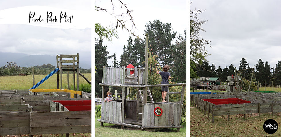 Children play on a wooden ship structure in a grassy park with maze-like paths. Trees and mountains are visible in the cloudy background.