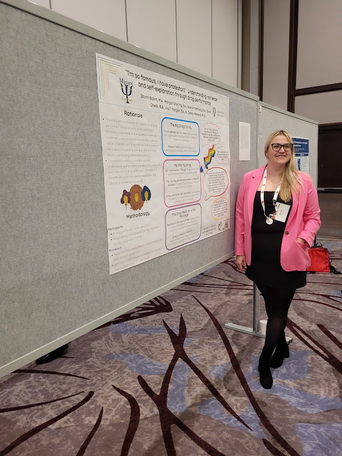 A young woman in a pink blazer stands in front of a conference poster