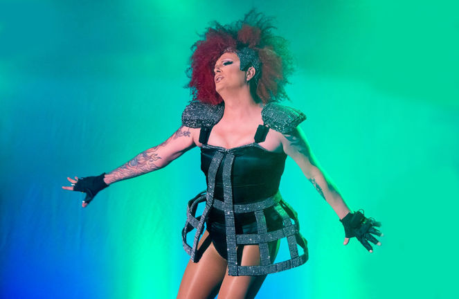 Person trägt ein großes, herzförmiges Blumenarrangement aus rosa und orangefarbenen Blüten auf dem Kopf und einen leuchtend grünen Federumhang