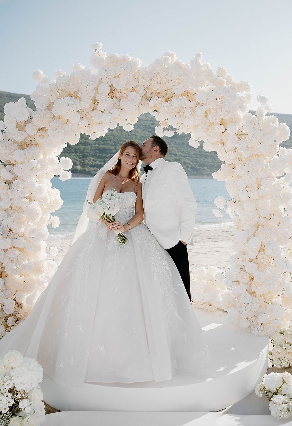 One and Only wedding. Bride in a white gown and groom in white tux, under floral arch by a beach. Groom kisses bride's head, both smiling. Sunny day.