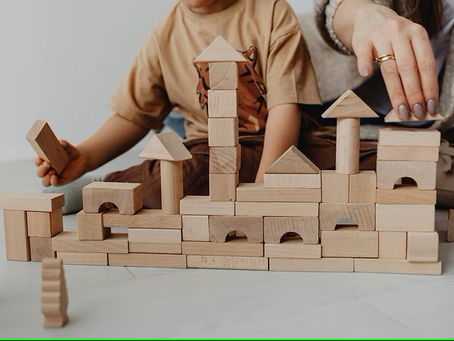 Child and adult hands building a tower with wooden blocks on a light floor. The child holds a block, and the mood is focused and playful.