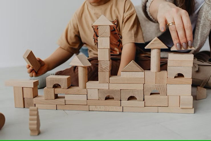 Child and adult hands building a tower with wooden blocks on a light floor. The child holds a block, and the mood is focused and playful.