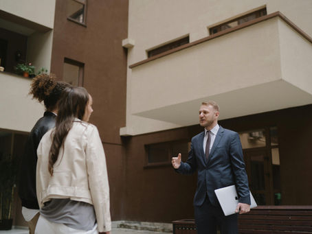 A professional real estate agent or consultant in a dark suit holding a laptop, gesturing while speaking to a couple outdoors. They are standing in front of a modern, multi-story residential building with neutral brown and beige tones, representing property management, real estate investment, or a housing consultation.