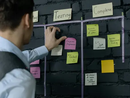 Person placing sticky note on a black brick wall with "Testing" and "Complete" sections. Notes are colorful; organized setting.