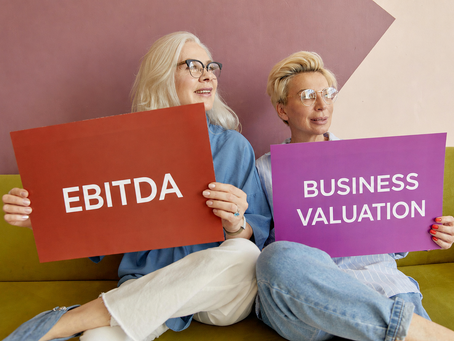 Two women holding colorful signs labeled “EBITDA” and “Business Valuation” while sitting together indoors, representing financial analysis, company valuation, entrepreneurship, business growth strategy, and corporate finance concepts.