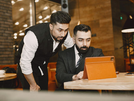 Two men focus on an orange tablet in a cozy café setting, with warm lighting and wood accents. One man is dressed in a suit, the other in a vest.