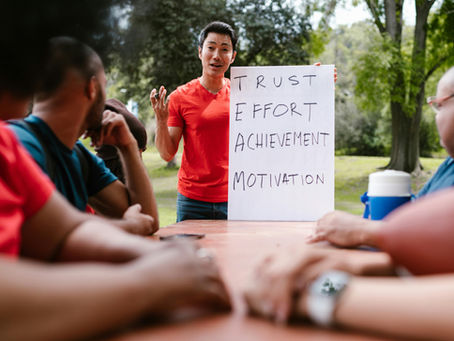 A person in a red shirt holds a sign reading "Trust, Effort, Achievement, Motivation" while speaking to a group at an outdoor table.