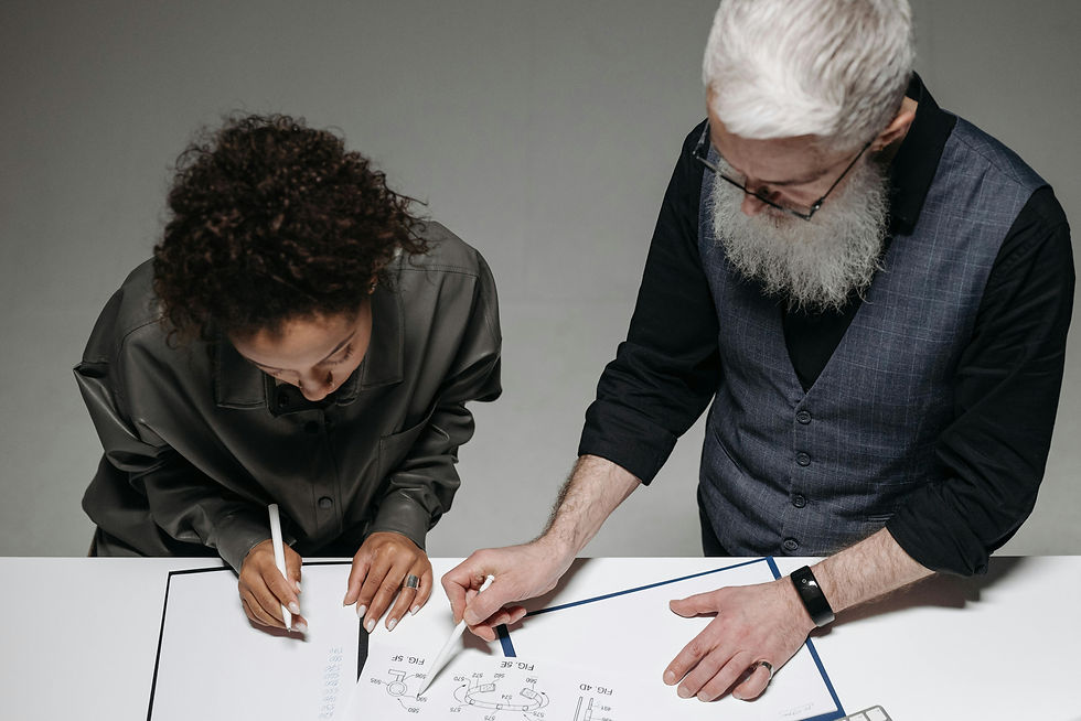 Two people are collaborating over a drawing on a table. One writes with a pen, the other points. Both focused. Gray, minimal setting.