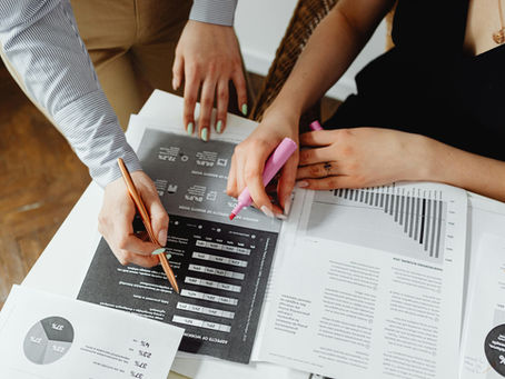 Hands analyzing documents with charts and graphs at a table. One holds a pink highlighter, another uses a pencil. Professional setting.