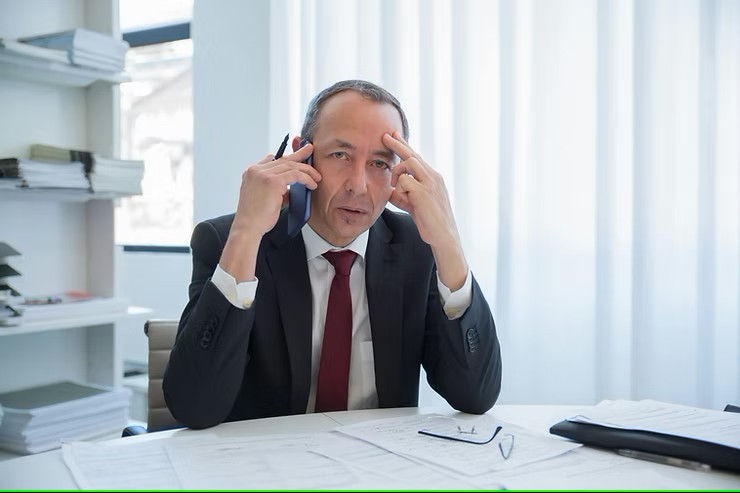 Man in a suit on phone, looking stressed, sits at a desk with papers. Light curtains in the background, bookshelves to the side.