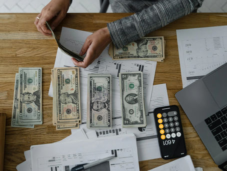 Hands count cash on a wooden table with documents, a laptop, and a smartphone calculator showing 4601. Bills are neatly organized.