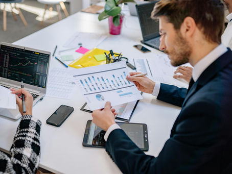 People in business attire analyze financial charts at a white desk with laptops, papers, and a potted plant in a well-lit office.