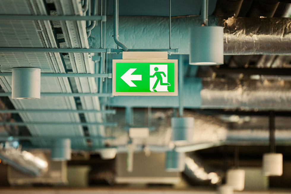 A green illuminated emergency exit sign hanging from an industrial ceiling, featuring a white running figure and a left-pointing arrow. The background shows exposed pipes, cable trays, and ventilation ducts in a modern commercial or industrial facility.