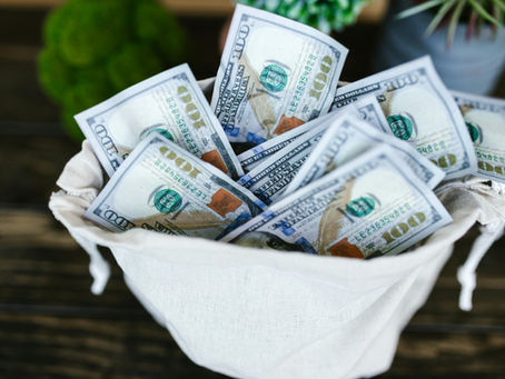 A beige sack overflowing with US $100 bills, set on a wooden surface. Green plants are blurred in the background, creating a rich mood.