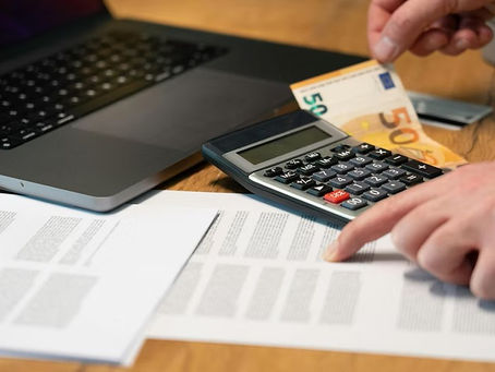 Hands hold a 50-euro note and use a calculator on a table with documents and a laptop. The setting is a work or financial environment.