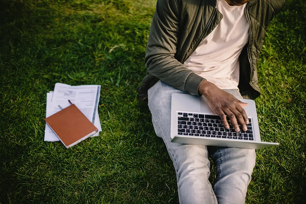 Person sitting on grass using a laptop, wearing a jacket and jeans. Notepad and papers lie nearby, in a relaxed outdoor setting.