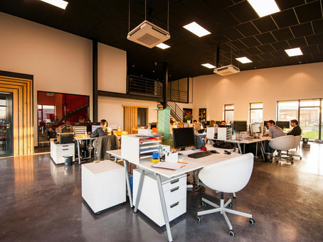 Modern office with people working at desks, computers on, white furniture, and large windows. Bright lighting and open design create a busy yet inviting atmosphere.