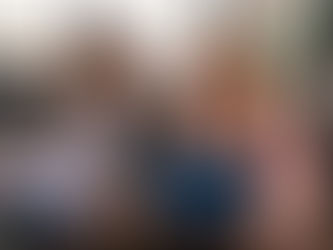 Two women in business attire discuss documents at a table in an office setting. One wears gray plaid, the other pink. They're focused and engaged.