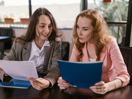 Two women in business attire discuss documents at a table in an office setting. One wears gray plaid, the other pink. They're focused and engaged.