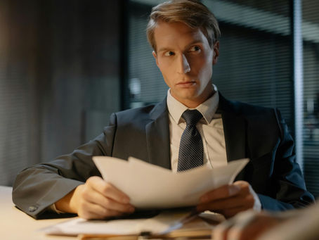 A medium, eye-level shot captures a young man with blonde hair, dressed in a professional black suit, white shirt, and patterned navy tie. He is seated at a desk in a dimly lit office, holding a stack of white papers in both hands. His expression is serious and alert as he looks off-camera to his right, as if listening intently or reacting to something in a meeting. The background is dark with office blinds, and a warm, low light source illuminates the desk and his face, creating a moody, cinematic atmosphere of professional scrutiny or interrogation.