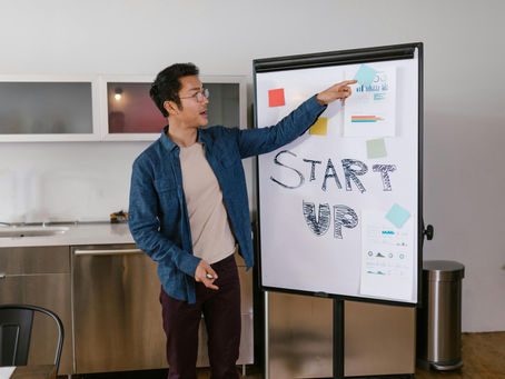 An Asian man in a blue denim shirt and glasses gestures toward a whiteboard during a business presentation. The board features the words "START UP" in bold, hand-drawn letters, along with several data charts and colorful sticky notes. The setting is a modern office or kitchen workspace.