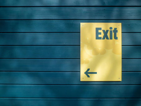 An outdoor photograph, featuring a yellow sign reading "EXIT" in black text with an arrow pointing left below it, affixed to blue-green wooden planks. Dappled shadows fall across the frame, partially obscuring the left side of the sign.