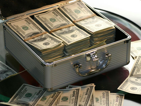 A metal briefcase filled with stacks of U.S. 100-dollar bills on a glass table, surrounded by more scattered bills.