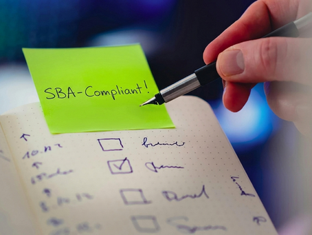 Hand holding a pen over a notebook checklist with "SBA-Compliant!" on a green sticky note. The background is blurred.