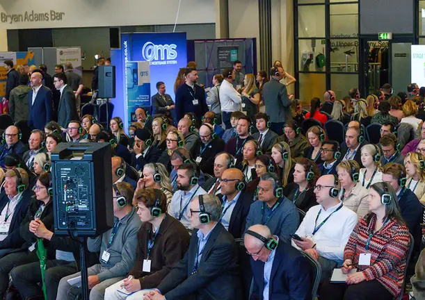 A packed audience wearing wireless headphones during a Stage Two session at the Building Societies Show 2026, held at the Coventry Building Society Arena, with the OMS sponsor stand visible in the background.