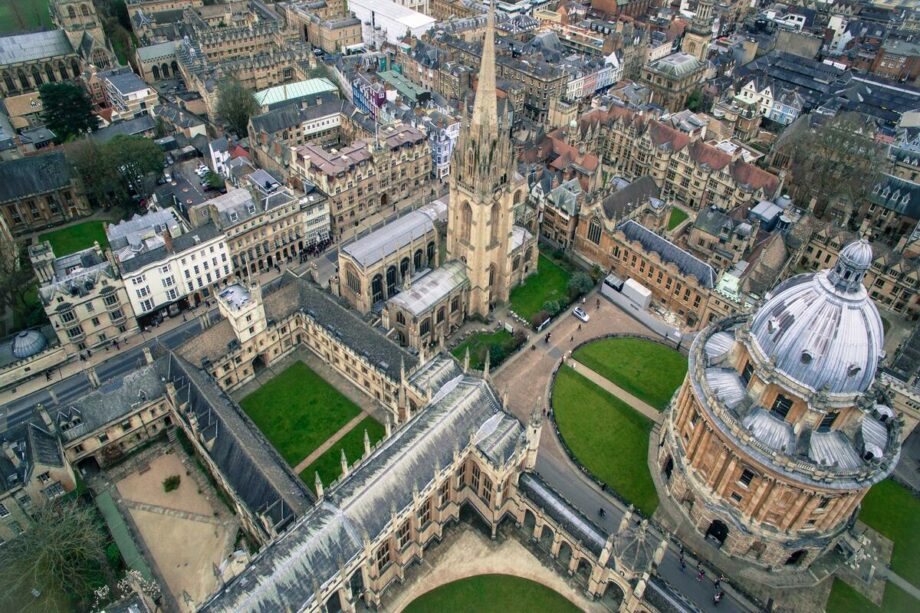 Aerial view of Oxford, showcasing historic stone buildings with spires and a dome, surrounded by greenery and a bustling cityscape.