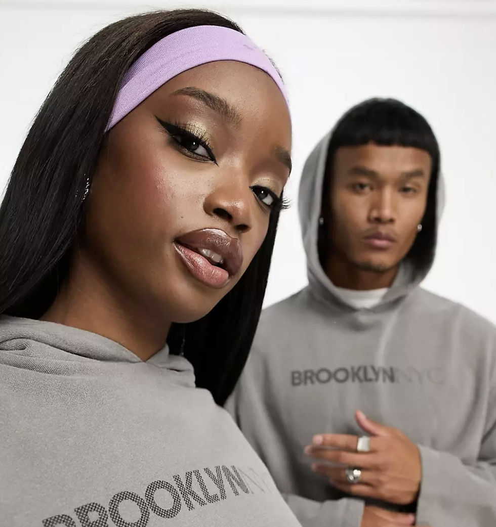 Two people in gray hoodies with "BROOKLYN NYC" text. Woman in foreground with a lavender headband, both exude a confident mood. White background.
