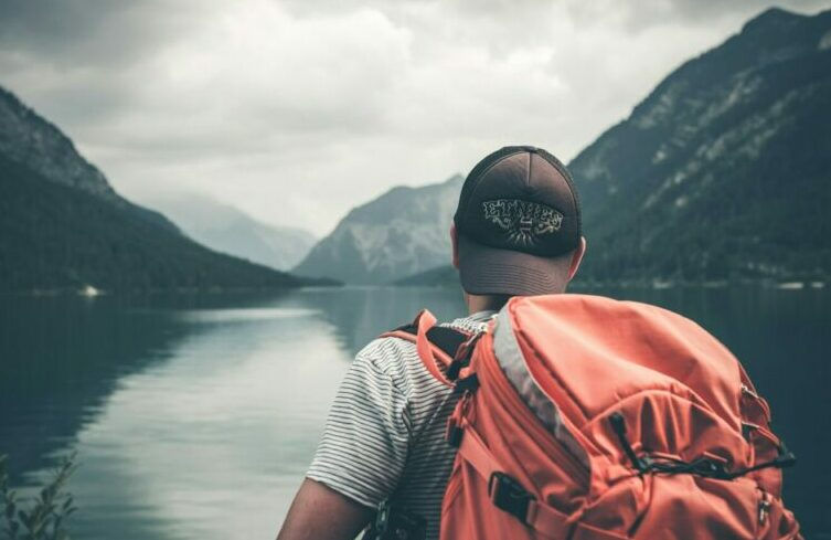 Man staring at a lake wearing a backwards baseball cap and carrying a large backpack on his back