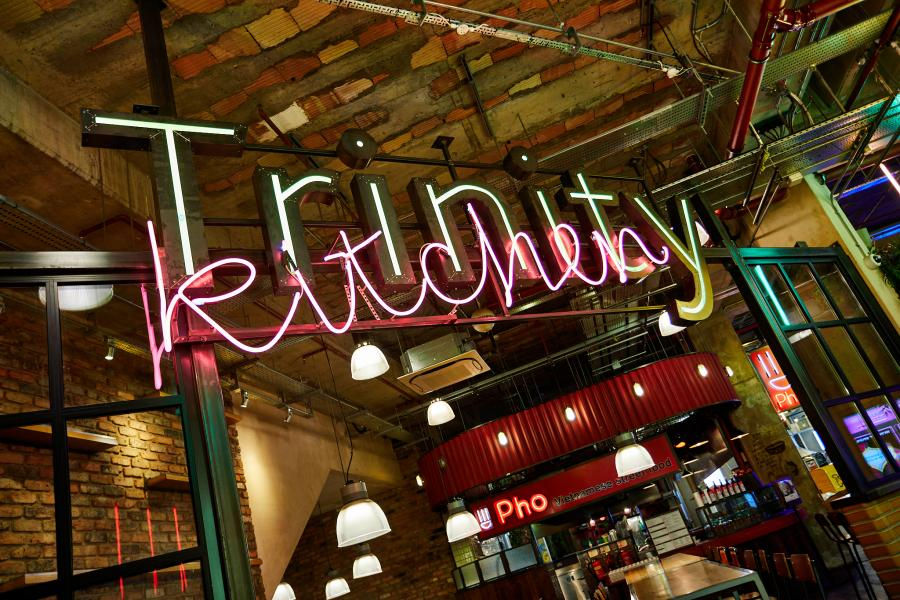 Neon signs reading "Trinity Kitchen" hang in an industrial-style food court. Brick walls and pendant lights add to the vibrant atmosphere.
