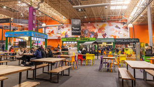 Indoor market hall with colorful chairs, people dining, and stalls like "Mr Mackerel" and "Istanbul Bakery." Vibrant mural in background.