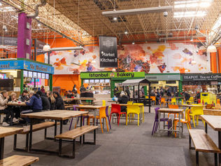 Indoor market hall with colorful chairs, people dining, and stalls like "Mr Mackerel" and "Istanbul Bakery." Vibrant mural in background.