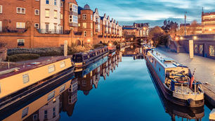 Canal at dusk with narrowboats reflecting in the water. Red brick buildings line the banks, and the sky is a serene blue, creating a peaceful mood.