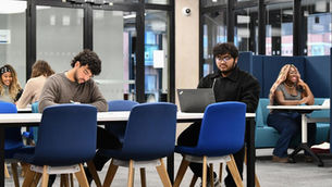 People are studying in a modern library with blue chairs. One is writing, another on a laptop, while others chat in the background.