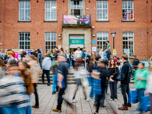 Students bustling outside a red brick building with a "Welcome to LUU" banner. Sign reads "Leeds University Union." Motion blur adds energy.