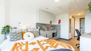 Modern studio apartment with a bed covered in a patterned orange and white blanket. Kitchen area with gray cabinets. Bright and cozy mood.
