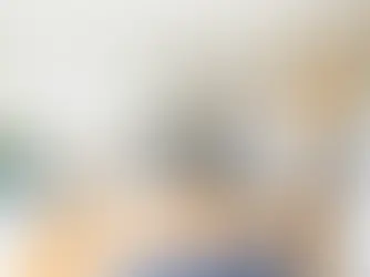 Modern studio apartment with a bed covered in a patterned orange and white blanket. Kitchen area with gray cabinets. Bright and cozy mood.