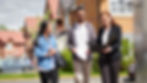 Real estate agent showing documents to a couple in a suburban neighborhood. The couple appears attentive, houses in the background.