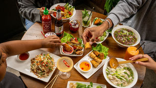 People enjoying a Vietnamese meal at a wooden table, featuring pho, spring rolls, salad, and drinks. Bright, colorful, and lively setting.